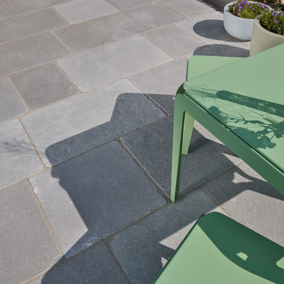 Terrasse mit grauen Kalksteinplatten, grünem Metalltisch und Blumentöpfen im Sonnenlicht.
