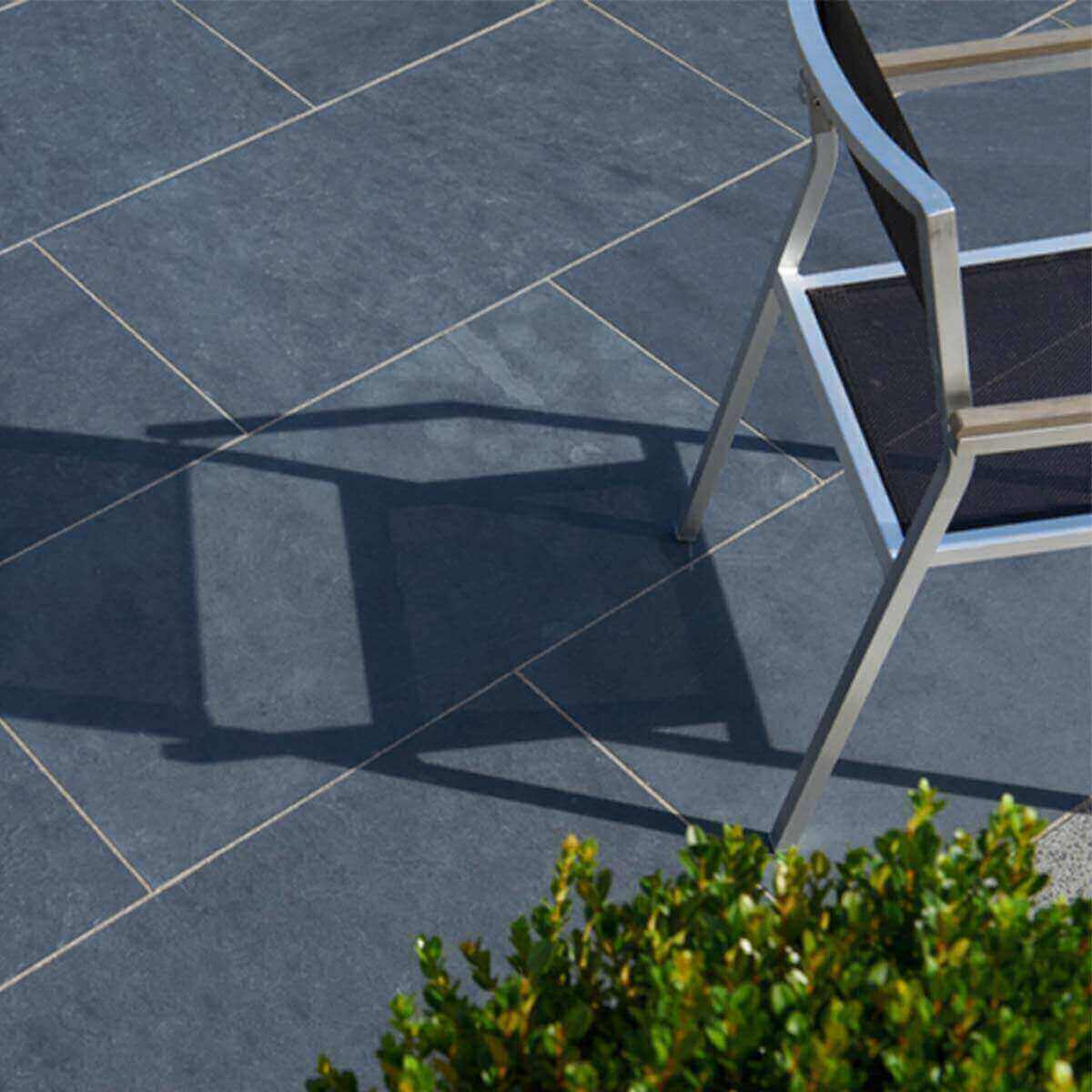 Terrasse mit dunklen Schieferplatten und modernem Metallstuhl neben einer grünen Pflanze.“