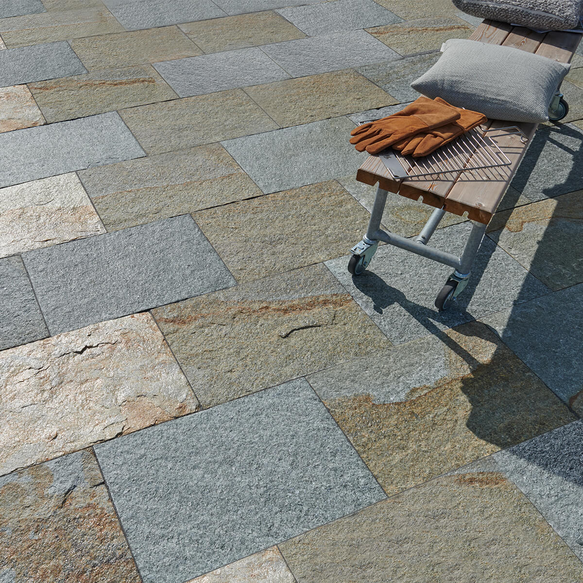 Terrasse mit mehrfarbigen Gneisplatten in Grau- und Brauntönen, daneben eine Holzbank mit Kissen und Grillhandschuhen.