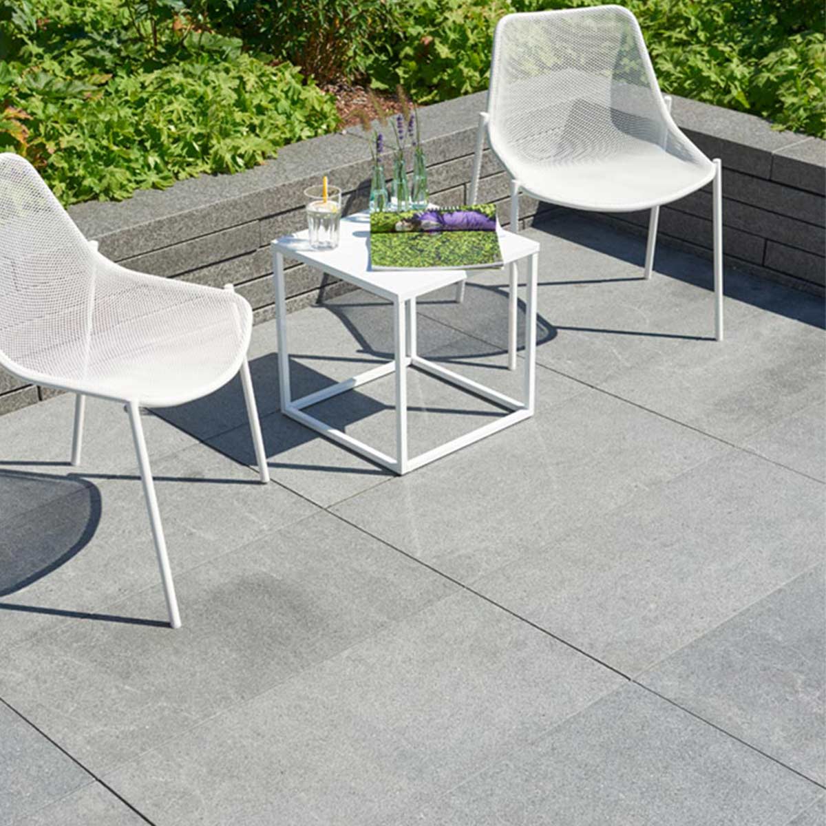 Moderne Terrasse mit grauen Granitplatten, weißen Stühlen und kleinem Tisch vor einer bepflanzten Mauer.