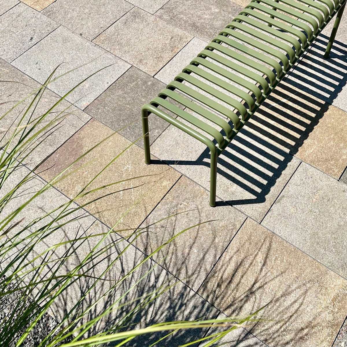 Terrassenfläche aus mehrfarbigen Porphyrplatten mit grüner Metallbank und Ziergräsern.