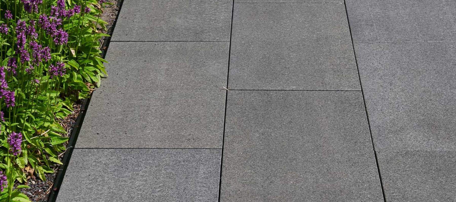 Graue Granit-Terrassenplatten neben einem Beet mit violett blühenden Pflanzen.
