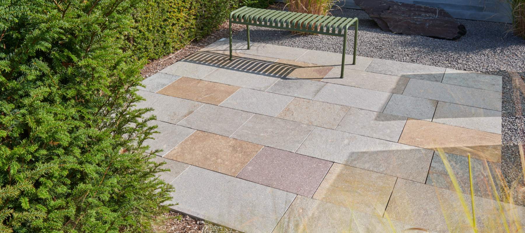 Terrasse mit mehrfarbigen Porphyrplatten in Grau-, Braun- und Rottönen sowie einer grünen Metallbank im Garten