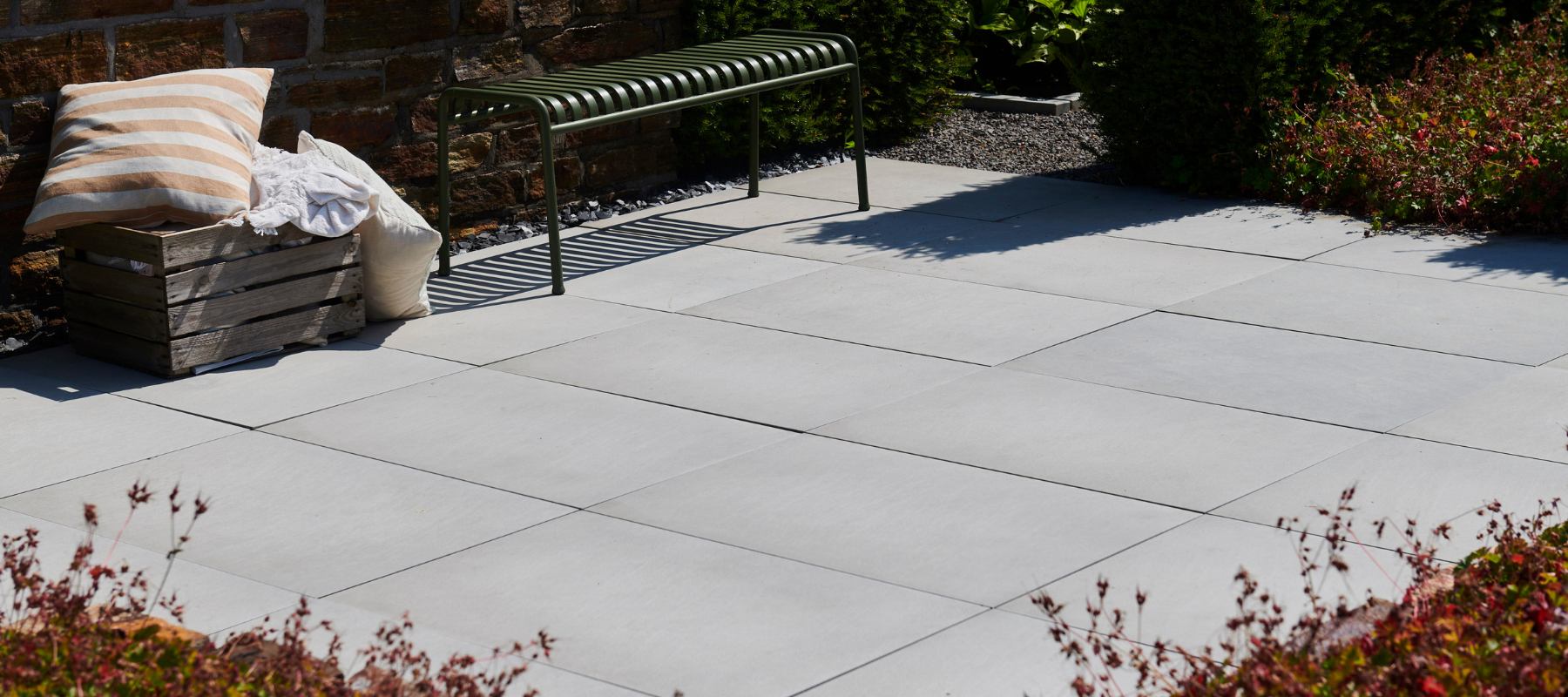 Terrasse mit hellgrauen Sandsteinplatten, grüner Metallbank und Holzkiste mit Kissen vor einer Natursteinmauer.